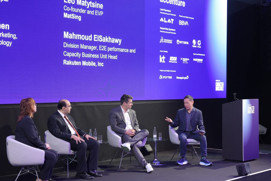 Four people are seated on stage in a panel discussion at the MWC25 Sports and Entertainment Summit. They sit in modern chairs against a backdrop showcasing names and logos. The person on the far right gestures animatedly, while bottles of water rest on the floor at this professional conference.