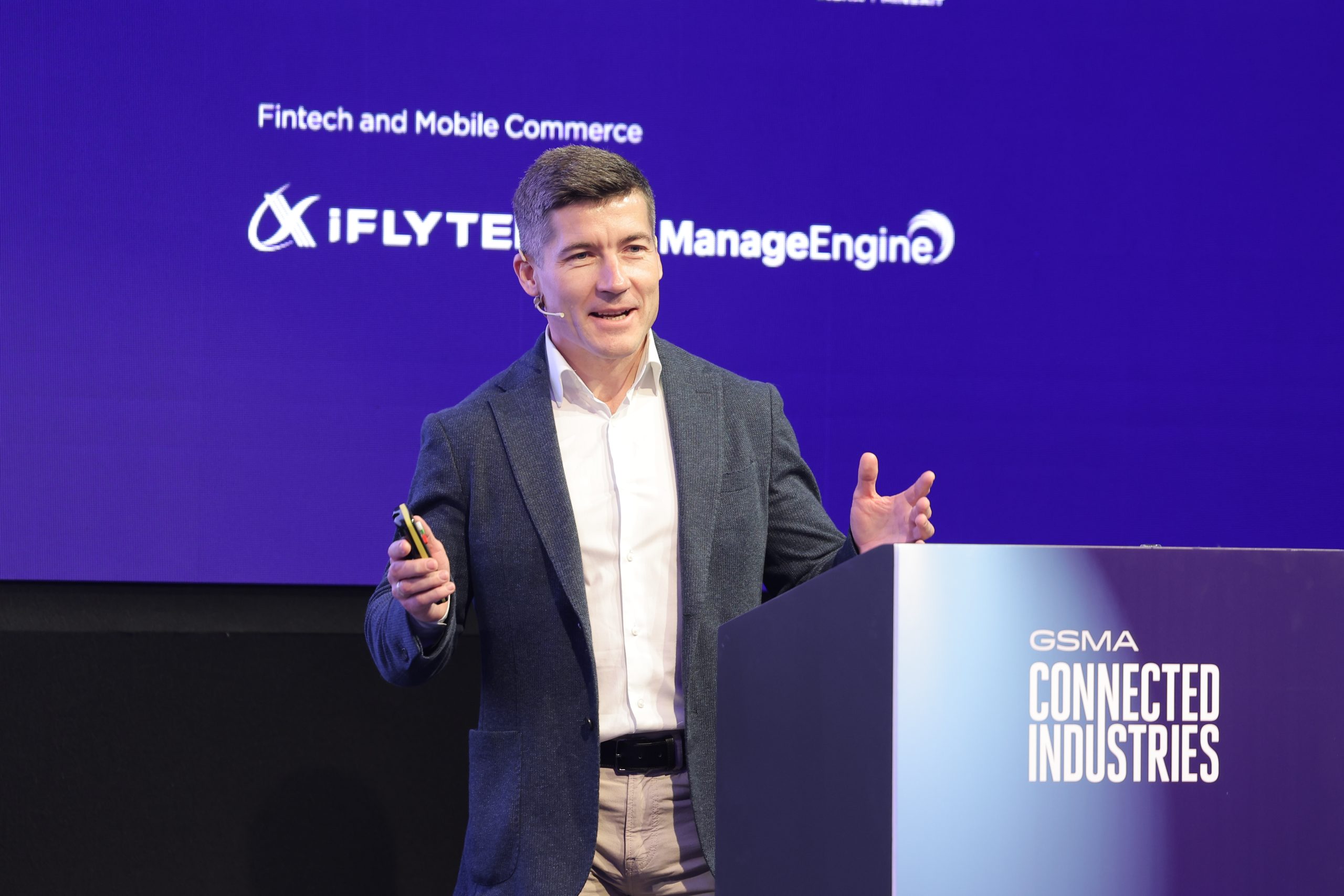 A person in a gray blazer and white shirt speaks on stage at the GSMA Connected Industries event. They stand at a podium with the event name, addressing the audience. The backdrop is blue with logos for iFlytek and ManageEngine, and text reading "Fintech and Mobile Commerce.