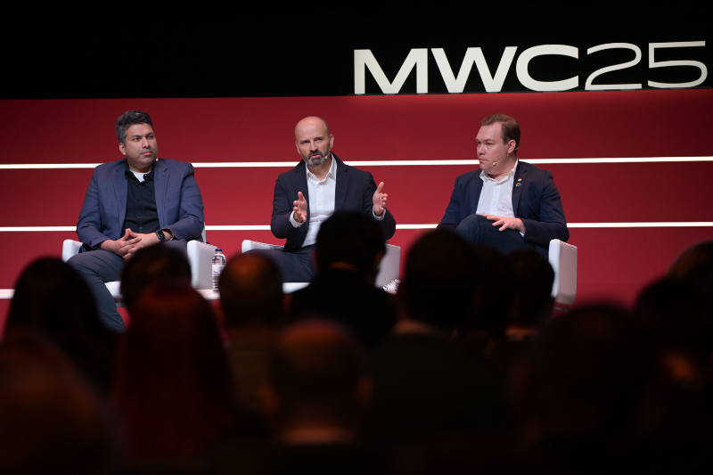 Three men in suits sit on a stage panel. The middle speaker gestures with both hands while talking. A large "MWC25" sign is visible behind them on a red backdrop. Audience members are seated in the foreground, watching the panel discussion.