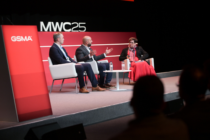 Three people are engaged in a panel discussion on stage at MWC25. They are seated in white chairs with a small table holding water bottles between them. The stage backdrop features the GSMA logo. The scene is professionally lit, and an audience is partially visible in the foreground.