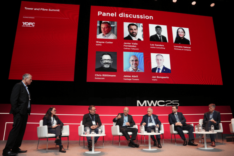 Panel discussion at the Tower and Fibre Summit stage featuring seven seated panelists and a standing moderator. The backdrop displays the names and photos of eight speakers, though one is not on stage. The event branding "MWC25" is visible on the set.