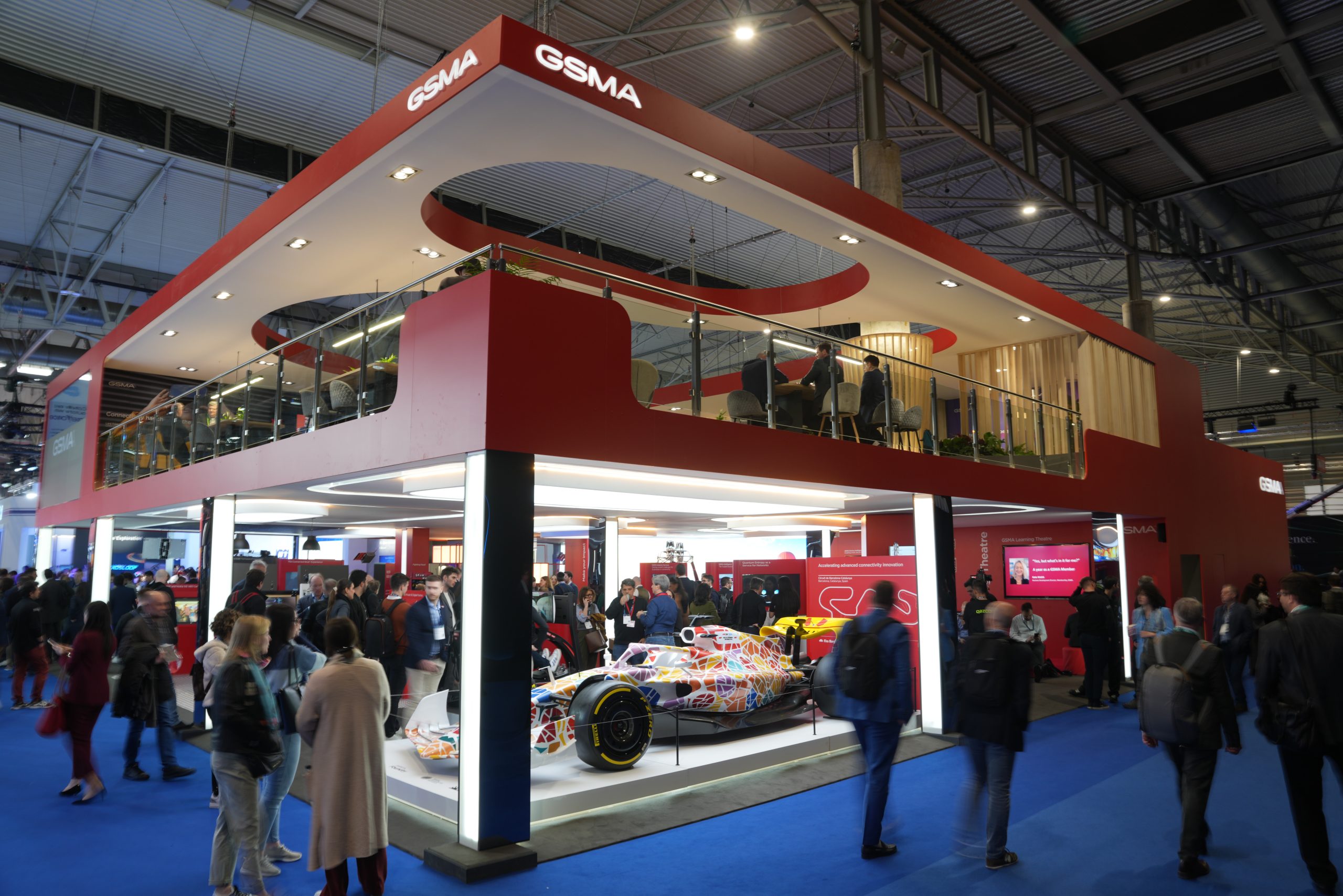 Exhibition booth with red and white design, displaying a colorful Formula 1 car. The booth has an upper level with people seated, and "GSMA" branding is visible. Attendees are walking and interacting on the blue-carpeted floor in a large convention hall.