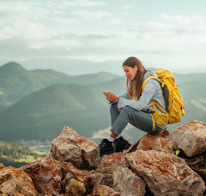 A young woman wearing a yellow backpack, blue jeans, and a light jacket sits on rocky terrain atop a mountain, looking at her phone. Misty mountains and a cloudy sky create a scenic background. She appears relaxed and focused.