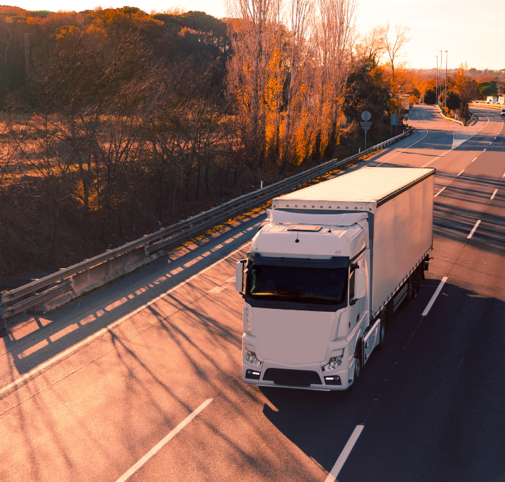 A white semi-truck with a large trailer drives along an empty, sunlit highway bordered by autumn trees and casting long shadows on the road. The scene is peaceful with clear skies and warm, golden light.