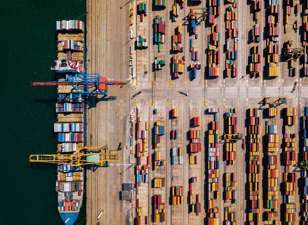 Aerial view of a busy container port with colorful shipping containers arranged in neat rows along piers. Two large cranes are positioned over a cargo ship docked at the pier. The water is visible on the left, contrasting with the industrial area.
