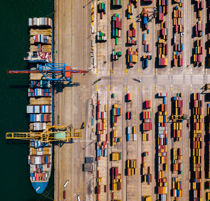Aerial view of a port with a large cargo ship docked on the left, loaded with colorful shipping containers. On the right, rows of stacked containers fill the yard, separated by wide concrete lanes. Green water is visible to the left.