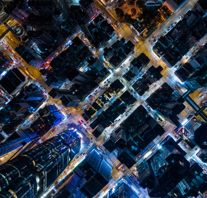 Aerial nighttime view of a city grid with illuminated streets forming a crisscross pattern. Skyscrapers and buildings cast dark silhouettes, while neon and streetlights create vibrant blue, yellow, and orange glows highlighting the urban landscape.