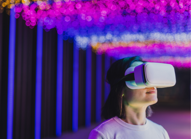 A person wearing a white virtual reality headset stands under a canopy of colorful, blurred purple, pink, and blue lights. The lights create a dreamy atmosphere, and the person appears immersed in the VR experience, wearing a white shirt and standing indoors.