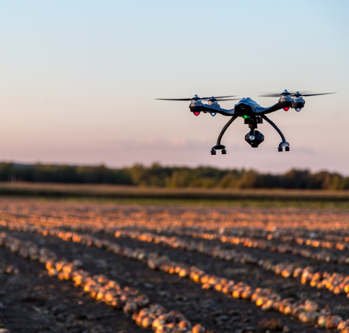 A black quadcopter drone hovers above a plowed farm field at sunset. The sky is clear with a soft gradient from blue to pink. Rows of crops or soil furrows stretch into the distance, bordered by a line of green trees in the background.
