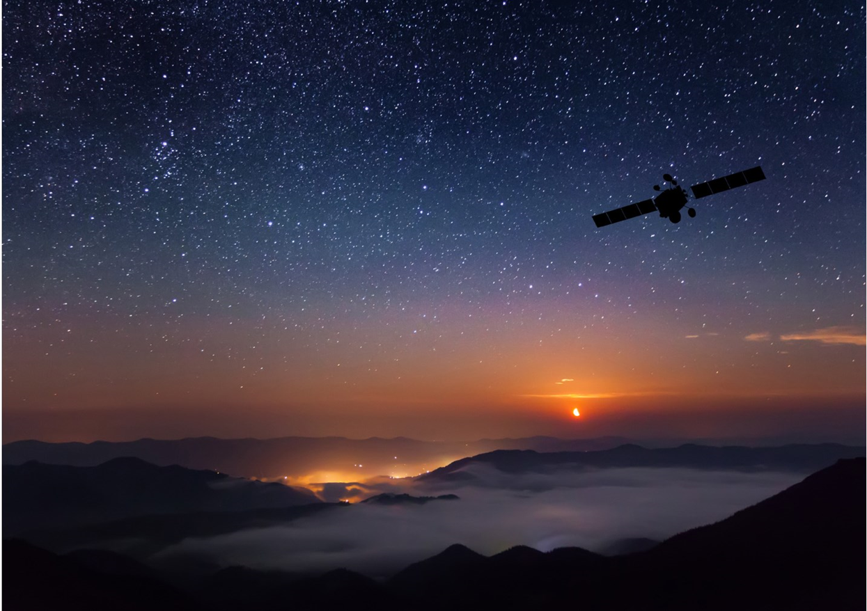A silhouette of a satellite orbits above mist-covered mountains at dusk. Distant city lights glow on the horizon as a setting sun and a sky full of stars create a dramatic, atmospheric scene.