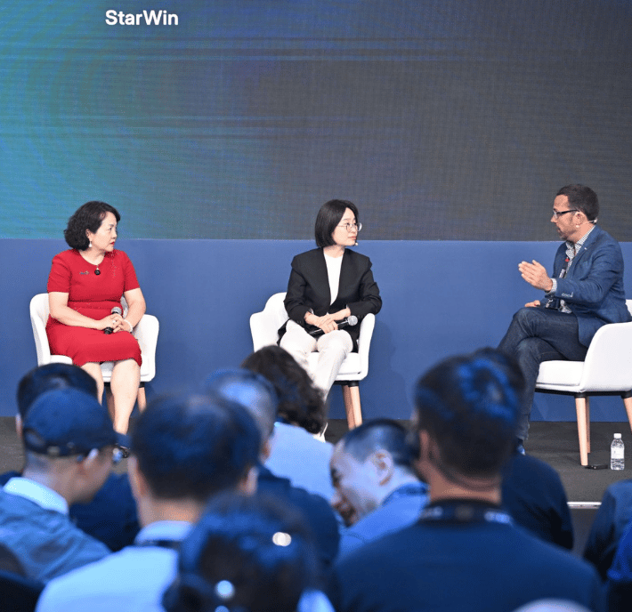 Three people sit on stage in white chairs, engaged in a panel discussion at a conference. A woman in a red dress, a woman in a black blazer, and a man in a jacket and jeans talk while the audience listens. A large screen behind them displays the word "StarWin.