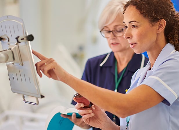 Two female nurses in uniforms are standing together in a hospital room. One points at a screen on a medical workstation while holding a smartphone, and the other looks on attentively. Both appear focused and engaged in reviewing patient information.
