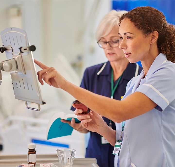Two nurses in light and dark blue uniforms review information on a digital tablet mounted on a stand. One nurse points at the screen while holding a medicine bottle; the other holds a notebook. Medical supplies and a hospital bed are visible in the background.
