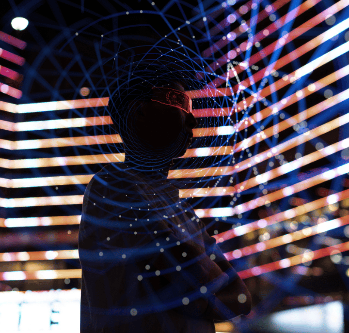Silhouette of a person wearing virtual reality goggles stands with arms crossed, surrounded by colourful, geometric, and swirling digital light patterns and grids, suggesting a futuristic, high-tech, or immersive digital environment.