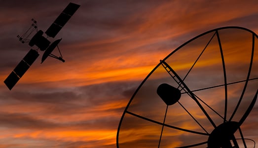 A satellite and a large satellite dish are silhouetted against a dramatic sunset sky streaked with orange and purple clouds, symbolizing communication technology and space exploration.