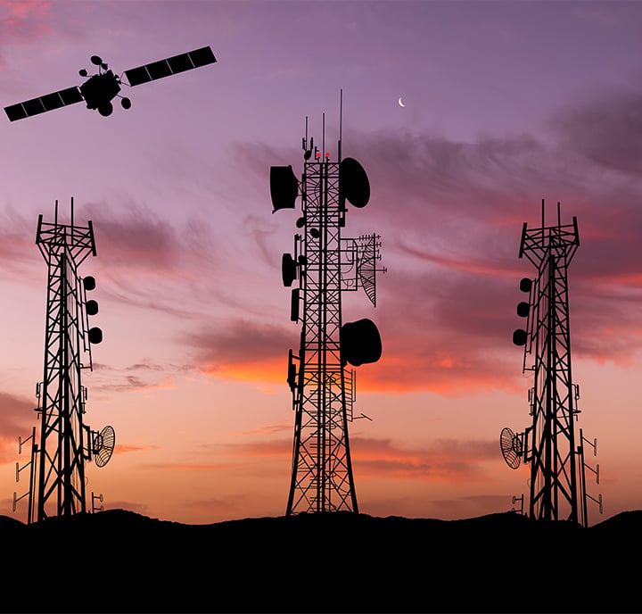 Silhouettes of three large telecommunications masts with various aerials stand on a ridge at sunset. A satellite is visible in the sky, and a crescent moon appears above wispy, colourful clouds in the background.