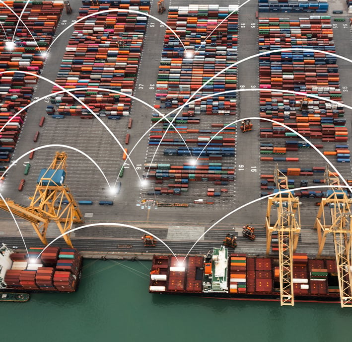 Aerial view of a busy shipping port with stacked colourful cargo containers, large yellow cranes loading containers onto ships, and white curved digital lines connecting various points, representing digital logistics or smart port technology.