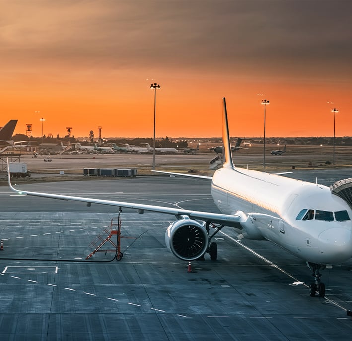 A white commercial aeroplane is parked at an airport gate during sunset. The sky is orange and gold, with the sun low on the horizon. Airport lights and other aeroplanes are visible in the background, and the aircraft is connected to a passenger boarding bridge.