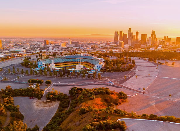 Aerial view of Dodger Stadium surrounded by empty car parks, with lush green trees in the foreground and the downtown Los Angeles skyline in the background under a warm, orange sunset sky.