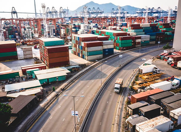 An aerial view of a busy shipping port with numerous colourful stacked cargo containers, lorries on curving roads, warehouses, and cranes in the background. Mountains and a clear sky are visible in the distance.