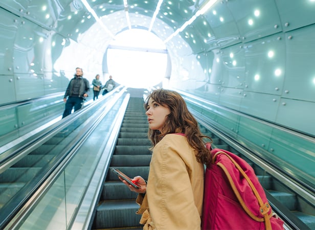 A young woman with brown hair, wearing a tan coat and carrying a pink rucksack, stands on an escalator holding her mobile. She looks over her shoulder as she ascends in a modern, brightly lit tunnel with people on adjacent escalators.
