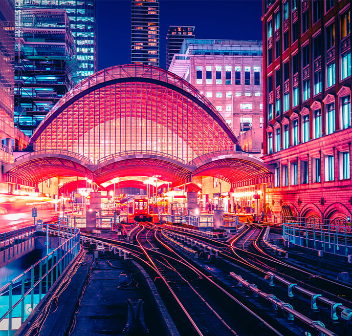 A vibrant, futuristic train station at night glows with bright neon pink and purple lights. Multiple rail tracks curve into the illuminated, arched glass canopy. Skyscrapers and city buildings surround the station, reflecting vivid colors. Light trails suggest moving trains.