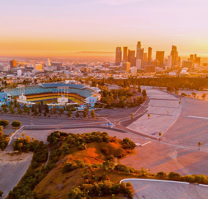 Aerial view of Dodger Stadium surrounded by empty car parks at sunset, with central Los Angeles skyscrapers in the background under an orange sky. Foreground includes trees and rolling hills bathed in golden light.