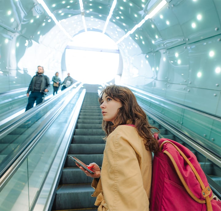 A young woman with long brown hair, wearing a tan coat and pink rucksack, stands on a moving walkway in a futuristic tunnel with glowing lights. She holds a mobile phone and looks upwards, while several people ascend on the opposite side.