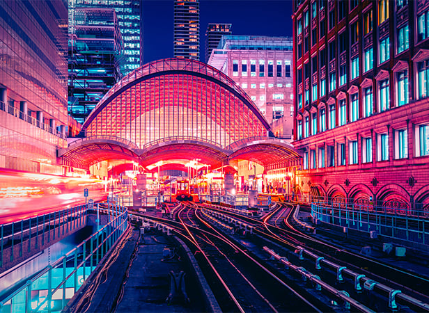 Vibrant night scene of a city railway station with arched glass roofing, illuminated in pink and purple lights. Multiple railway tracks lead towards the station. Surrounding modern and historic buildings are lit by colourful urban lighting.