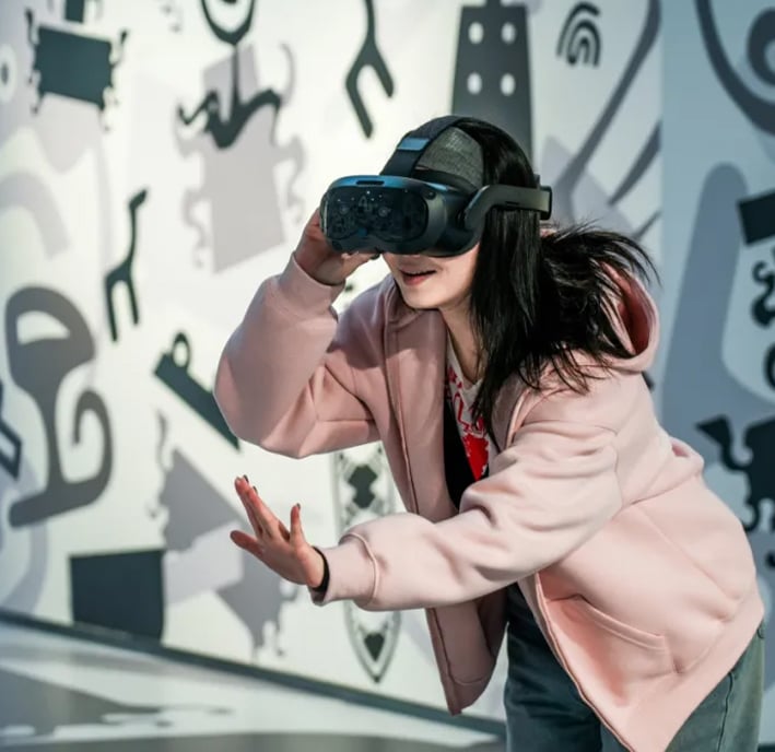 A young woman wearing a pink hoodie and a VR headset extends her hand forwards, interacting with a virtual environment. The background features large, abstract black-and-white graphics and playful shapes on the walls.