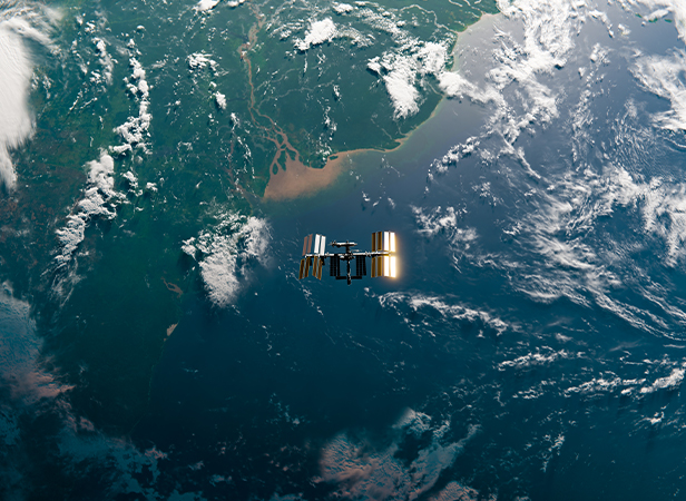 A satellite with reflective solar panels orbits above Earth, with continents, clouds, and the ocean visible below. Sunlight highlights land and water features, while the atmosphere curves along the planet’s edge.