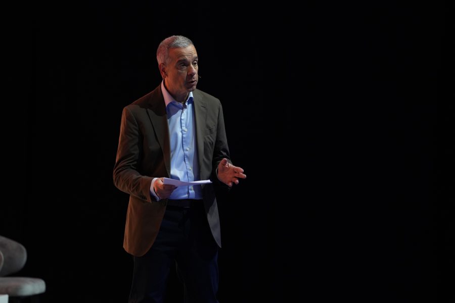 A man in a brown blazer, light blue shirt, and dark trousers stands on a dimly lit stage holding papers. He gestures with one hand whilst speaking, with a serious expression. The background is mostly dark, emphasising the figure.