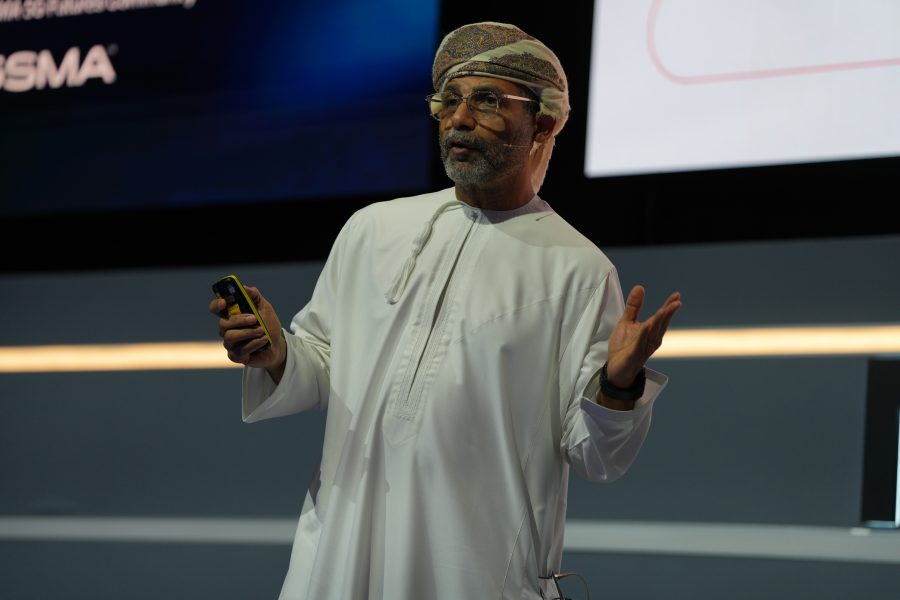A man wearing a traditional white Middle Eastern thobe and patterned head covering stands on stage, holding a yellow mobile and gesturing with his other hand, appearing to speak or present at an event. Dark and light screens are visible in the background.