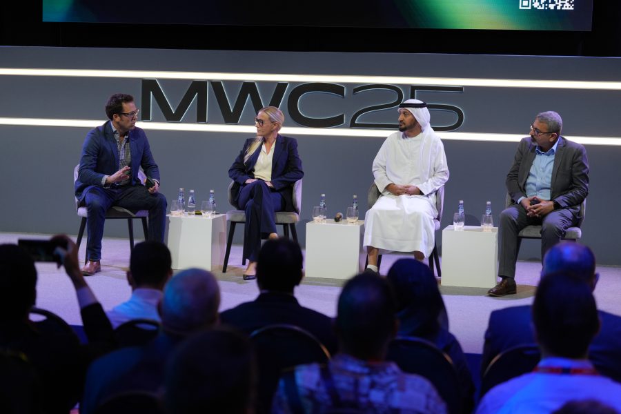 Four panellists, three men and one woman, sit on a stage in discussion at MWC25. They are seated on modern white chairs with water bottles in front of them, facing an audience. A large "MWC25" sign is illuminated behind them.