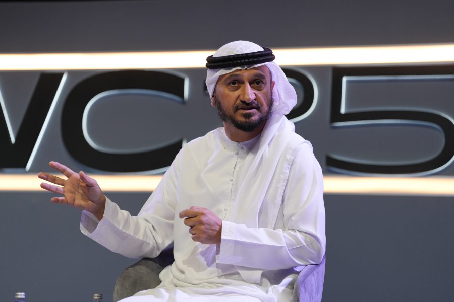 A man in a white kandura and black agal gestures while speaking on stage. He sits on a grey chair against a dark background with large, partially visible letters and yellow horizontal lights behind him.