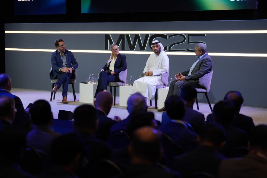 Four men in business attire sit on stage having a panel discussion at a conference. Behind them is a large "MWC25" sign. The audience is visible in the foreground, listening attentively to the speakers.