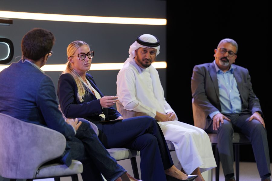 Four people sit on stage in modern chairs, engaged in a panel discussion. One woman gestures while speaking, flanked by three men, one of whom wears traditional Middle Eastern attire. All appear focused, with a dark background and horizontal light strips behind them.