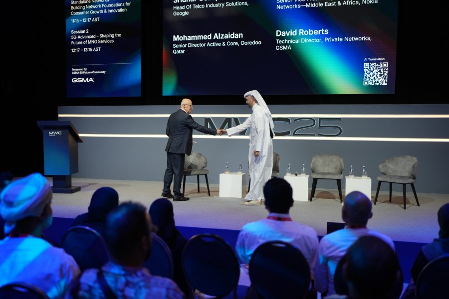 Two men shake hands on stage at a conference, with audience members seated in the foreground. Behind them, large screens display session and speaker details, including names: Mohammed Alzaidan and David Roberts. Four empty chairs are arranged on stage.