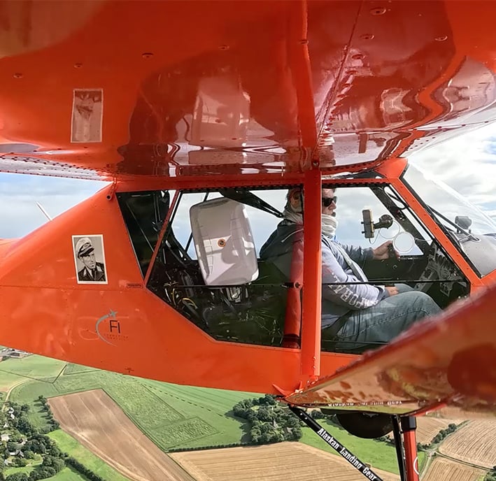 Person piloting a bright orange small aeroplane over green and brown fields under a partly cloudy sky.