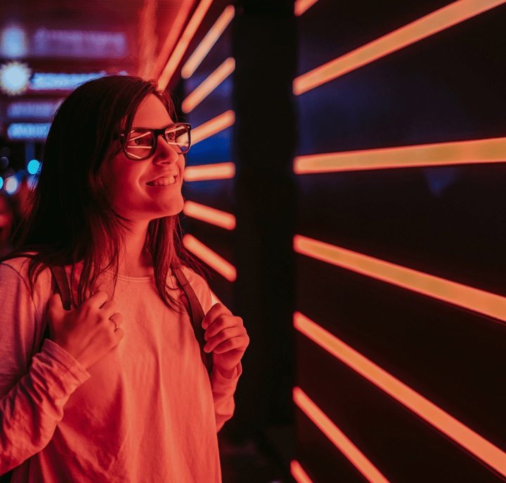 Woman with glasses smiling and standing by a wall with horizontal red neon lights at night, capturing the vibrant energy found in GSMA networks.