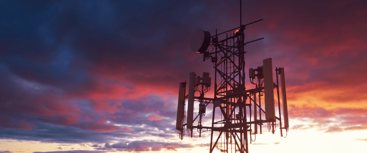 A silhouette of a tower with antennas stands against a dramatic sunset sky, filled with vibrant hues of orange, red, purple, and blue clouds. The sun is low on the horizon, casting a warm glow around the tower’s structure.