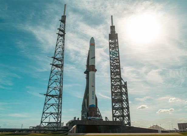 A rocket stands on a launch pad between two towers under a bright, partly cloudy sky.