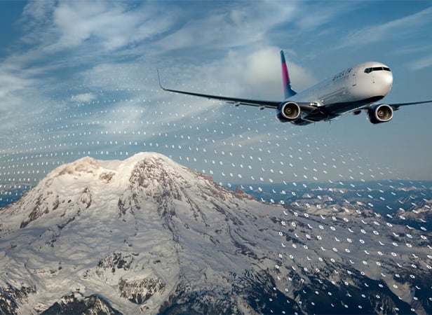 A commercial aeroplane flies over a snow-covered mountain under a blue sky with graphic wind patterns visible.