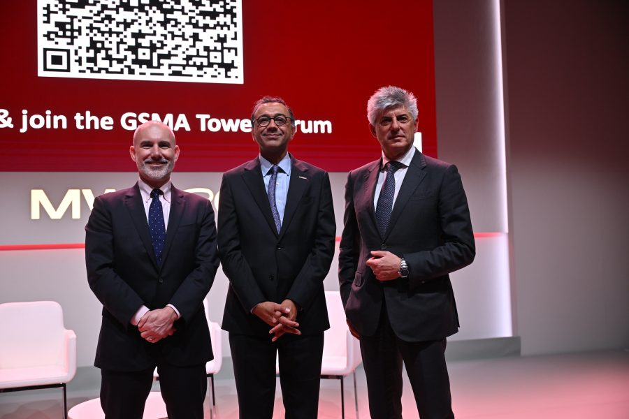 Three men in suits standing on a stage in front of a red screen with a large QR code and empty chairs.