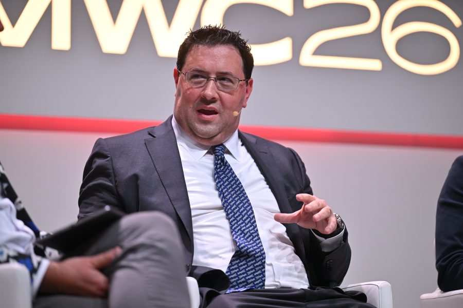A man in a suit and tie speaks while seated onstage at a conference with "MWC26" in the background.