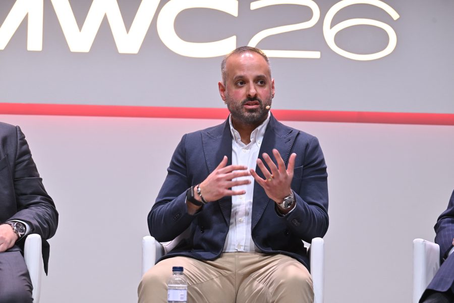 A man in a suit speaks on stage at an event, gesturing with his hands. "MWC 26" is visible behind him.