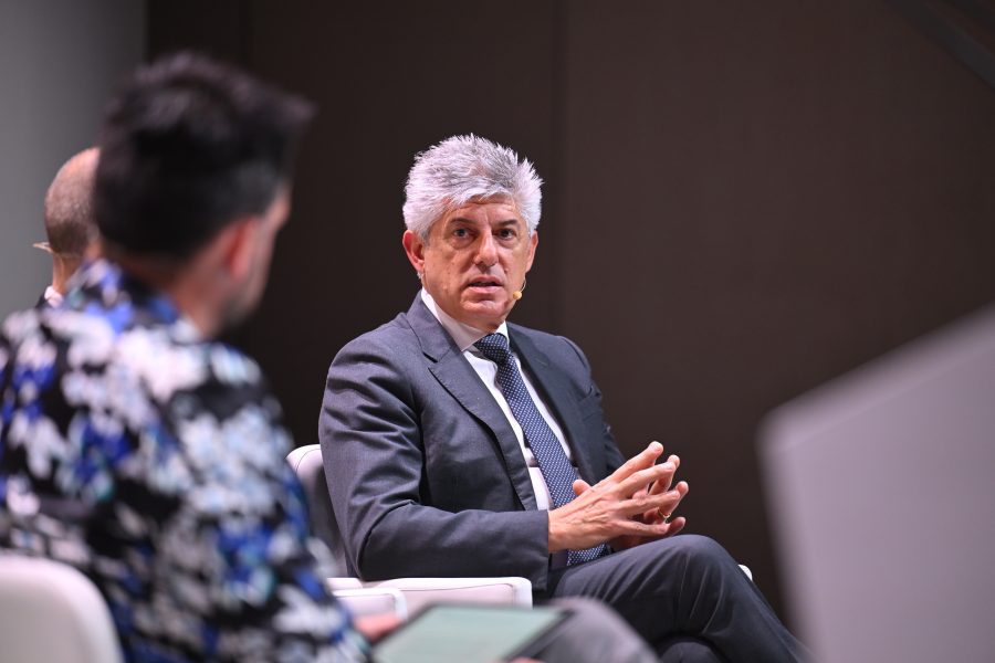A man in a suit speaks while seated on a panel, another person in focus listens in the foreground.