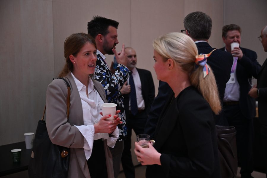 Two women talk at a networking event while others mingle in the background, holding drinks.