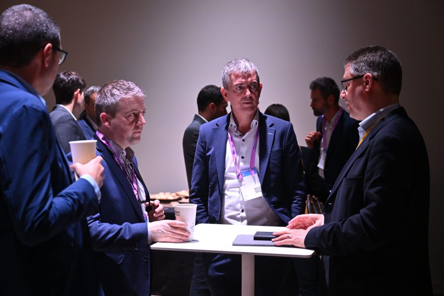 Four men in business attire stand around a high table, talking and holding cups at an event.