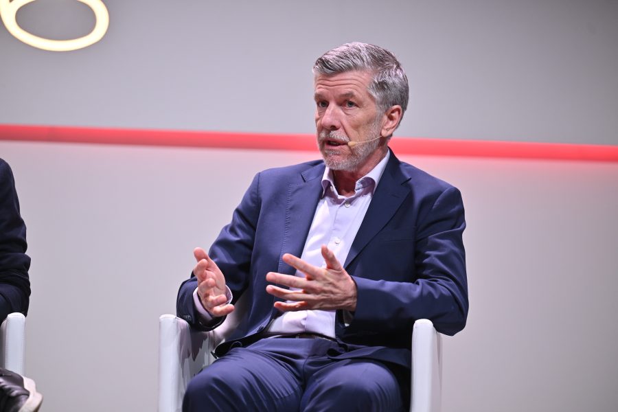 Man in a blue suit speaking with hand gestures, wearing a headset microphone, seated on a white chair.